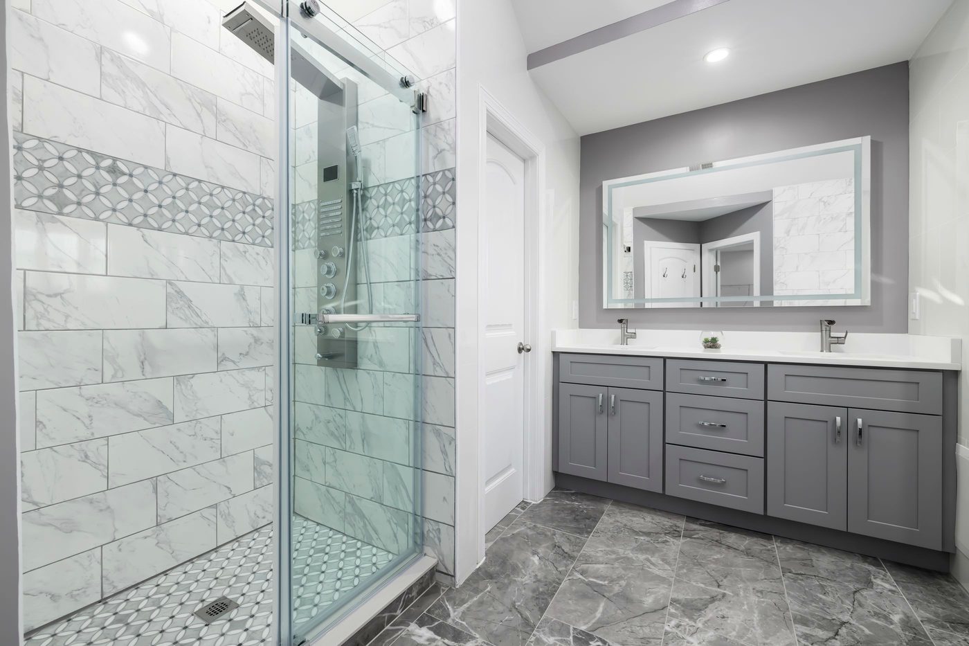 A finished bathroom with a tiled walk-in shower behind glass, a grey vanity with stone top, and a large mirror.