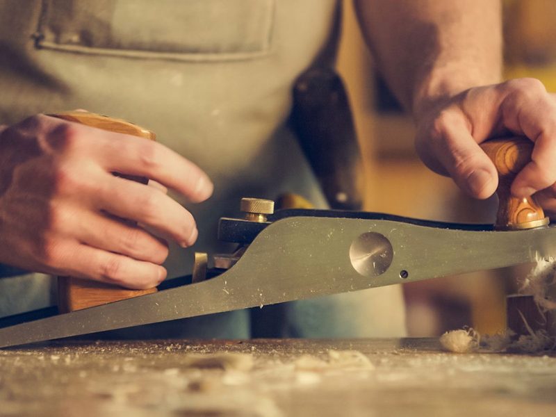 Hands running a hand plane along a length of timber, curls of shaving lifting off.