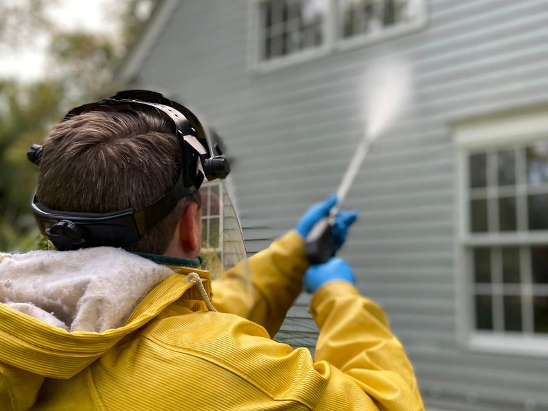 A worker in protective gear cleaning house siding with a pressure washer.