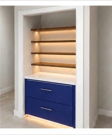 A custom built-in pantry cabinet in a white recess: four floating wood shelves lit by concealed LED strips above a white quartz counter, with two navy soft-close drawers below.