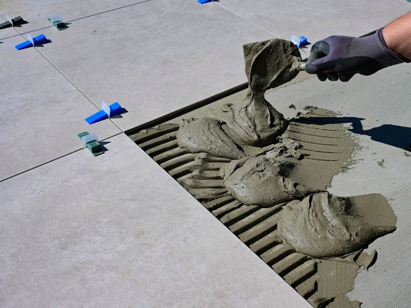 A gloved hand combing mortar with a notched trowel and setting floor tile with levelling spacers.